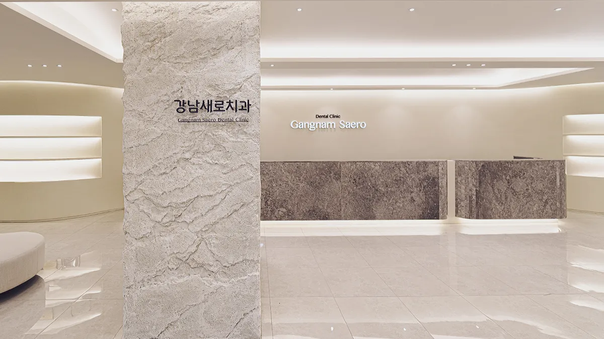 강남새로치과의원 로비, '강남새로치과' 글자가 붙은 거친 질감의 흰 석재 기둥 뒤로 회갈색 대리석 접수대와 'Gangnam Saero' 간판이 보이는 모습